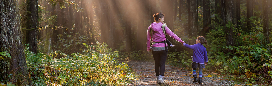 A woman walking through a forest, holding hands with a child and carrying a baby in a carrier