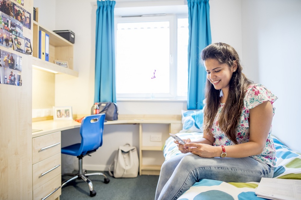 Female student sat on a bed in a room with a desk and chair