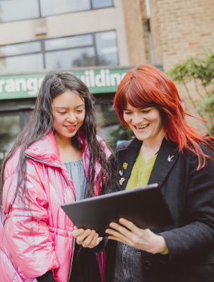 Two students standing outside looking at table, smiling